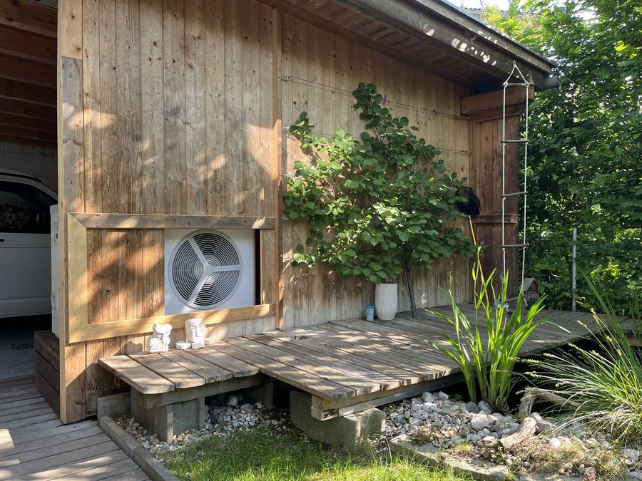 Wärmepumpen-Außeneinheit in eine Holzfassade eingelassen, davor eine Terrasse mit Teich