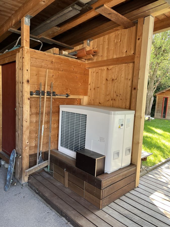 Wärmepumpen-Außeneinheit auf einem Holzpodest unter einem überdachten Vorbau
